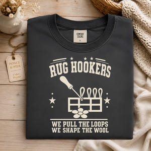 May include: A charcoal gray t-shirt featuring a cream-colored graphic with the text RUG HOOKERS, WE PULL THE LOOPS, WE SHAPE THE WOOL, and an illustration of a rug hooking tool and loops. The shirt has a Comfort Colors label indicating size L.