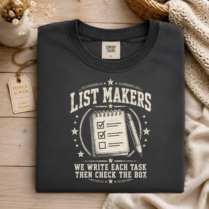 May include: A dark gray crew neck t-shirt featuring a white graphic of a notepad with a pencil and the text "LIST MAKERS" at the top and "WE WRITE EACH TASK THEN CHECK THE BOX" at the bottom, with a Comfort Colors size L label at the neck.