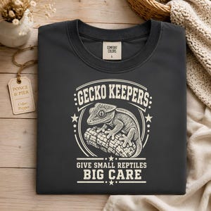 May include: A pepper-colored t-shirt featuring a cream-colored graphic of a gecko on a branch. The text reads GECKO KEEPERS GIVE SMALL REPTILES BIG CARE. The shirt has a Comfort Colors label and a tag labeled Ponce & Pier Color: Pepper.