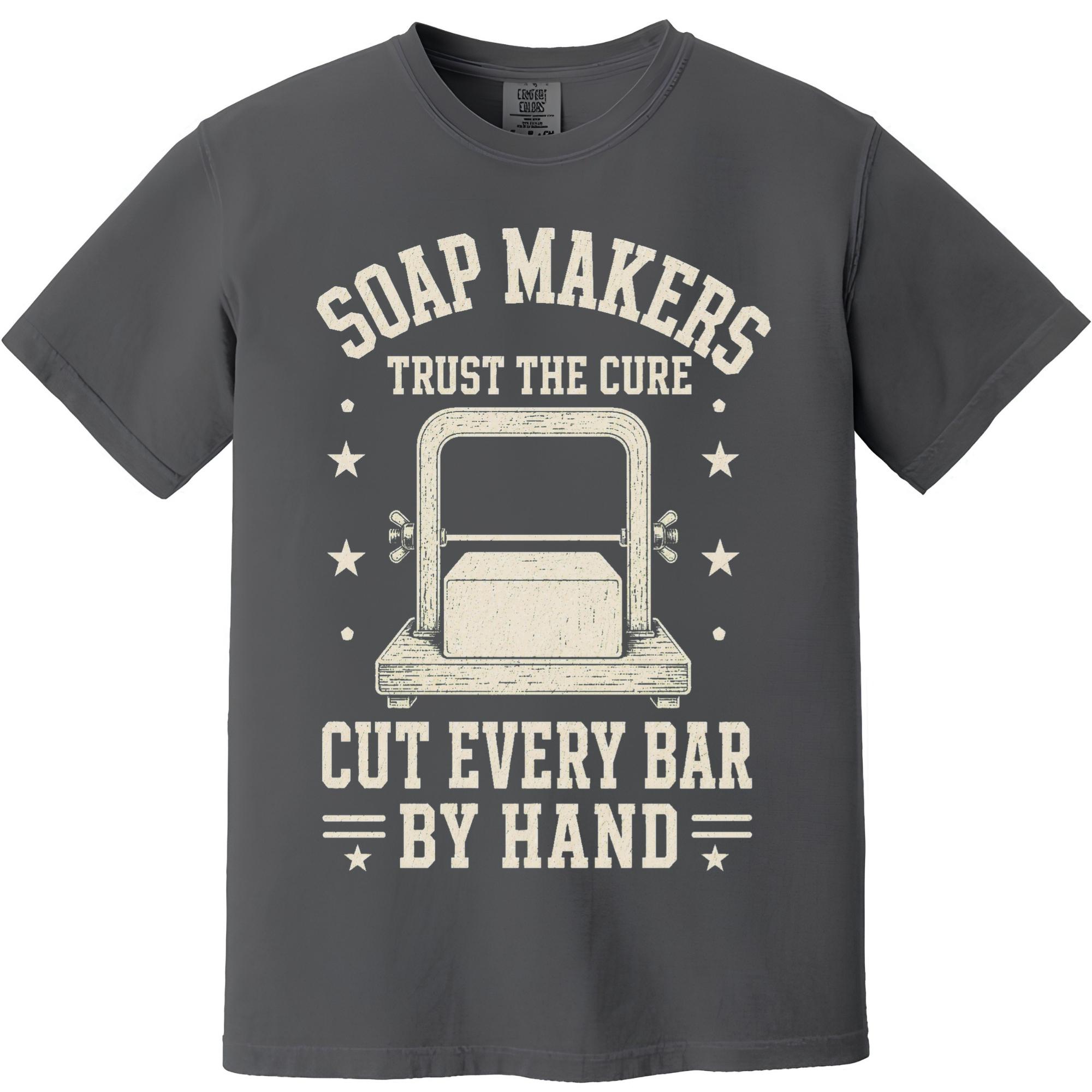 May include: A charcoal gray short-sleeve t-shirt featuring a cream-colored graphic of a soap cutter with a block of soap. The text reads: SOAP MAKERS TRUST THE CURE CUT EVERY BAR BY HAND, surrounded by small stars and horizontal lines.