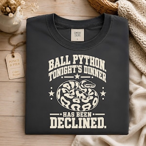May include: A charcoal gray t-shirt featuring a cream-colored graphic of a coiled ball python. The text reads: BALL PYTHON. TONIGHT'S DINNER HAS BEEN DECLINED. The shirt has a Comfort Colors label at the neck and a tag labeled Ponce & Pier.