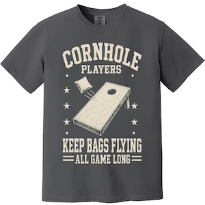 May include: A charcoal gray short-sleeve t-shirt featuring a cream-colored graphic of a cornhole board and a bean bag in flight. The text reads CORNHOLE PLAYERS KEEP BAGS FLYING ALL GAME LONG, accented by small stars and dots on both sides.