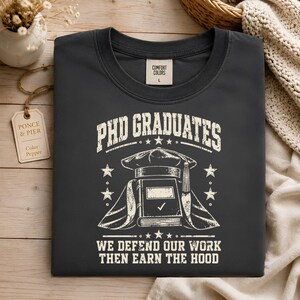May include: A dark gray t-shirt featuring a white graphic print. The text reads "PHD GRADUATES" and "WE DEFEND OUR WORK THEN EARN THE HOOD." The design includes a graduation cap, a book with a checkmark, and stars. A tag inside reads "COMFORT COLORS L."