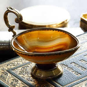 May include: A small, shallow bowl crafted from banded amber-colored agate, featuring a polished finish. The bowl is set on a circular brass base and includes a single, ornate, curved brass handle attached to the rim with a beaded metal edge.