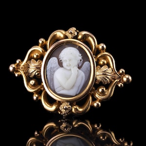 Puede incluir: Broche dorado con camafeo ovalado que presenta un querubín blanco con alas. El querubín está sobre un fondo oscuro y enmarcado por un borde dorado. El broche tiene un diseño de filigrana ornamentado con detalles florales y de cuentas.