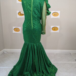 May include: A green sequined formal gown with a mermaid silhouette. The dress features a single long sleeve with gold accents and a large ruffled shoulder detail. A long green zip runs down the back.