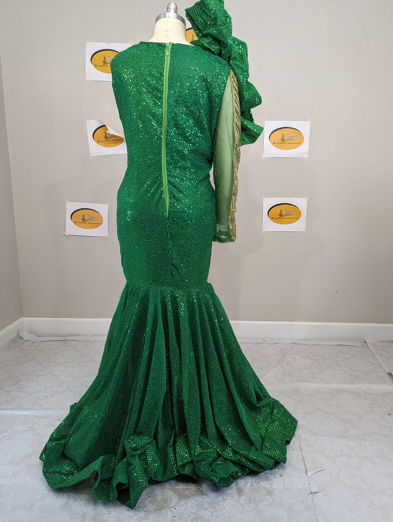May include: A green sequined formal gown with a mermaid silhouette. The dress features a single long sleeve with gold accents and a large ruffled shoulder detail. A long green zip runs down the back.