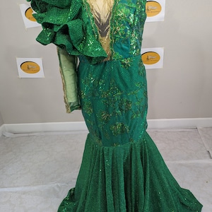 May include: A green sequined formal gown with a mermaid silhouette. The dress features a large ruffled sleeve, gold embellishments, and floral embroidery. The bodice has a sheer panel. The dress is displayed on a mannequin.