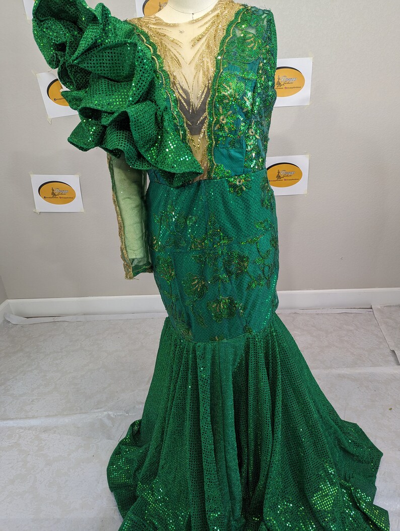 May include: A green sequined formal gown with a mermaid silhouette. The dress features a large ruffled sleeve, gold embellishments, and floral embroidery. The bodice has a sheer panel. The dress is displayed on a mannequin.