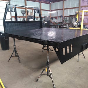以下が含まれることがあります： ダイヤモンドプレート模様の黒いスチール製トラック用フラットベッド。ヘッドエイクラックには切り抜きが施されています。フラットベッドは質感のある表面で、長方形のスロットがある垂直サイドパネルと中央のリアヒッチプレートを備え、金属製のジャッキスタンドで支えられています。