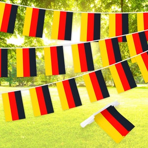 May include: A string of yellow, red, and black German flags hanging against a green, leafy background. The flags are strung together with white string.