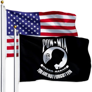 May include: A black POW MIA flag with a white silhouette of a soldier's head, a watchtower, and barbed wire. The flag reads "POW MIA You Are Not Forgotten". The flag is flying next to an American flag.