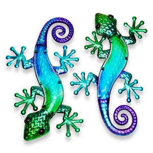 May include: Two metal gecko wall art pieces. The lizards are painted in shades of blue and green, with a curled tail. The gecko's feet are star-shaped. The eyes are black. The background is white.