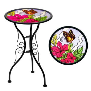 May include: A black metal side table with a round, glass top featuring a butterfly and floral design. The top has a black border and is supported by three curved legs. The table's design includes pink flowers, green leaves, and a butterfly.