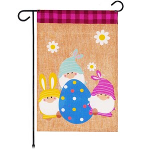 May include: A brown burlap garden flag with a pink and white checkered border. Three gnomes are holding a blue and white Easter egg with yellow, white, and pink polka dots. The gnomes are wearing blue, yellow, and pink hats.