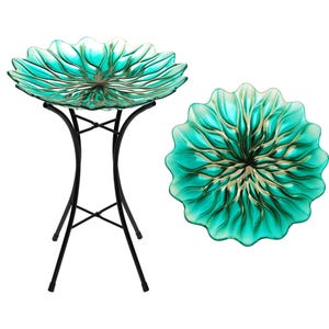 May include: A teal glass bird bath with a flower-like design, supported by a black metal stand. The bird bath has a scalloped edge and a gold-colored center. The stand has a tripod base.