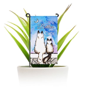 May include: A garden flag with a watercolor background featuring two cats sitting back to back. The flag has the text "Life is Better with Cats" on it.