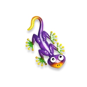 Könnte beinhalten: Ein Gecko-Wandschmuck aus Metall mit einem leuchtend violetten Körper mit weißen Sprenkeln, einem gelb gefärbten Schwanzende und hellgrünen Füßen. Das Tier hat große, runde weiße Augen mit schwarzen Pupillen und eine gelb-orange Schnauze.