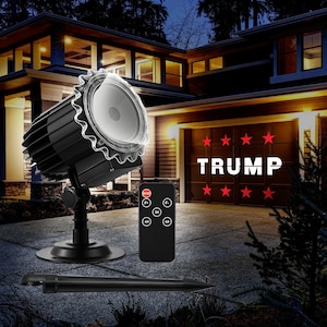 May include: A black projector with a clear lens projects the word "TRUMP" in white letters with red stars on a garage door. The projector is on a tripod and has a remote control.
