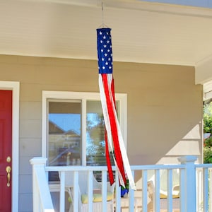 May include: A red, white, and blue American flag windsock hangs from the roof of a house with a white porch railing. The windsock is made of fabric and has a long, flowing tail.