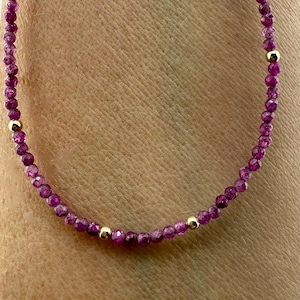 May include: A delicate necklace featuring small, faceted, deep pink gemstones strung together. The necklace is accented with small, gold-colored beads. The gemstones are a vibrant shade of pink, creating a beautiful contrast with the gold accents.