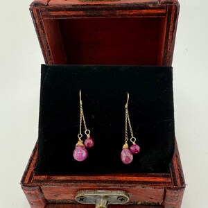 Puede incluir: Un par de pendientes dorados con piedras preciosas de color rubí, exhibidos dentro de un joyero de madera roja con interior de terciopelo negro. Los pendientes tienen un delicado diseño de cadena.