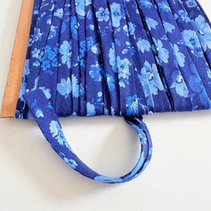 May include: Close-up of a blue floral fabric with a pleated design. The fabric features a pattern of light blue flowers on a darker blue background. A matching blue fabric handle is attached to the pleated fabric.
