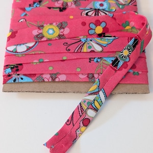 May include: A roll of pink bias binding featuring a floral and butterfly design in blue, yellow, and white. The tape is folded and ready for use, ideal for sewing and craft projects. The tape is measured in centimetres.