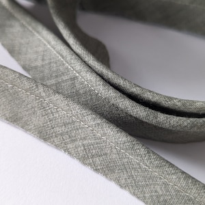 May include: Close-up of grey fabric bias tape with a white stitched edge. The tape is folded over and appears to be made of a woven material. The background is white.