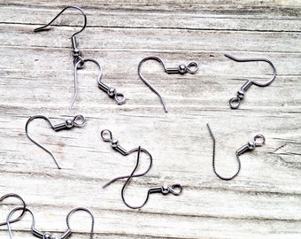 May include: A close-up of nine silver earring hooks on a light wood surface. The hooks are all the same size and shape, with a small loop at the top and a small ball at the bottom.