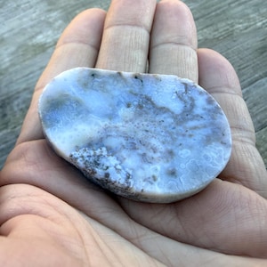 May include: A polished, oval-shaped stone with a light blue and white marbled pattern, featuring subtle brown and gray inclusions. The stone is held in an open hand, showcasing its natural beauty and unique texture. The stone is a decorative item.