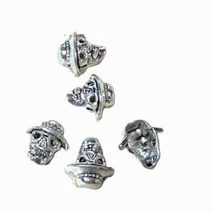 May include: Five silver-colored skull charms, each wearing a hat. The skulls have detailed facial features and are set against a white background. The hats have a decorative band around the crown.