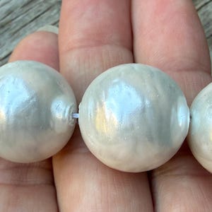 May include: A strand of large, round, white beads with a lustrous, pearlescent finish. The beads are strung together on a thin white cord, showcasing a smooth, reflective surface that catches the light, creating subtle highlights across each sphere.