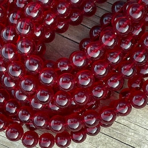 May include: Strands of deep red, translucent, round beads. The beads are strung together, creating a necklace or bracelet. The beads are smooth and shiny, reflecting light. The background is a wooden surface.
