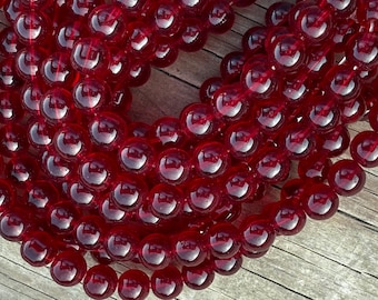 Puede incluir: Primer plano de hilos de cuentas redondas, translúcidas y de color rojo intenso. Las cuentas están ensartadas, posiblemente para un collar o pulsera. Las cuentas son lisas y brillantes. El fondo es una superficie de madera.