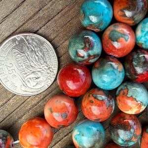 May include: A collection of colorful, round beads in shades of red, orange, teal, and green. A US quarter coin is placed next to the beads for size comparison. The beads have a marbled appearance.