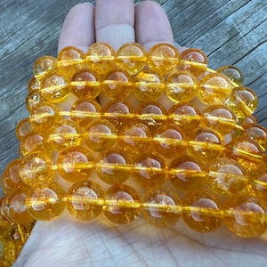 May include: A collection of golden-yellow, translucent beads strung together. The beads are round and appear to be made of a polished, glassy material. The beads are held in a hand, with a wooden surface in the background.