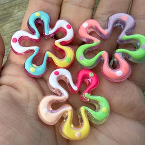 May include: Five colorful flower-shaped glass beads. Each bead features a unique combination of colors, including pink, green, yellow, blue, and purple, with white polka dots. The beads have a glossy finish and are displayed on a hand.
