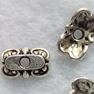 May include: Two silver metal bead caps with ornate designs. One cap is rectangular with a center hole, the other is a flower shape with a center hole.