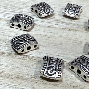 May include: Silver metal beads with a decorative design. The beads have three holes and are perfect for jewelry making.