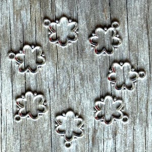 May include: Eight silver metal flower-shaped charms with a loop on each side. The charms are arranged on a wooden surface.