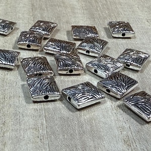 May include: A collection of silver-toned metal square beads with a textured design. The beads have a hole in the center for stringing.
