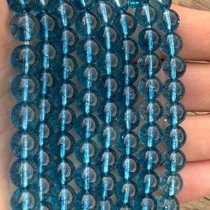 May include: Strands of translucent, round, blue-green beads. The beads are strung together, creating a textured, layered effect. The beads are a consistent size and shape, with a glossy finish. The overall color is a vibrant teal.