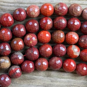 May include: Close-up of a strand of faceted red jasper gemstone beads. The beads are round with a geometric pattern, and vary in shades of red and brown. These beads are suitable for jewelry making.