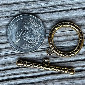 May include: Gold-toned metal toggle clasp with a round ring and a bar with two knobs. The clasp is shown next to a US quarter for size comparison.