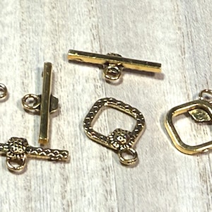 May include: Five gold-toned metal clasps for jewelry making. The clasps are in different shapes, including a square, a rectangle, and a bar. The clasps have decorative details, such as a floral design.