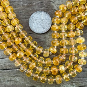 May include: Strands of faceted, golden-yellow gemstone beads. The beads are strung together with a silver-colored string. A silver coin with text is next to the beads. The beads are a warm, amber color and have a faceted cut.