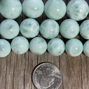 May include: Rows of pale green, round gemstone beads with subtle white marbling. A U.S. quarter coin is included for size reference. The beads have a smooth, polished surface and are arranged on a wooden surface.