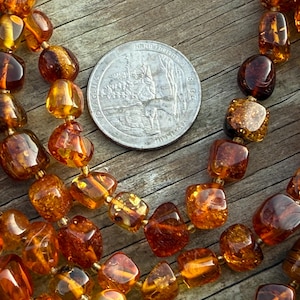 May include: Close-up of amber-colored beaded necklaces with a quarter coin for size comparison. The beads are various shapes and sizes, with a warm, translucent glow. The necklaces are strung on a gold-colored thread, and the coin shows a detailed design.