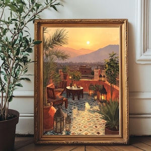 Op de afbeelding: Een ingelijste prent van een dakscène bij zonsondergang. Het kunstwerk toont een terras met een tafel, stoelen en lantaarns, omringd door potplanten. Het warme kleurenpalet omvat oranje, rood en blauw, met een bergketen op de achtergrond.
